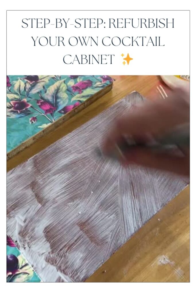 A hand applying white primer or paint on a wooden surface, surrounded by colorful floral fabric. The text at the top reads: "STEP-BY-STEP: REFURBISH YOUR OWN COCKTAIL CABINET ✨".