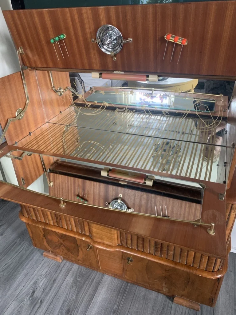 A vintage wooden display cabinet with glass shelves, mirrored back, and decorative brass accents. The cabinet has a partially open front, revealing interior shelves and multiple racks. The bottom section features cabinetry with intricate woodwork. It stands on a hardwood floor.