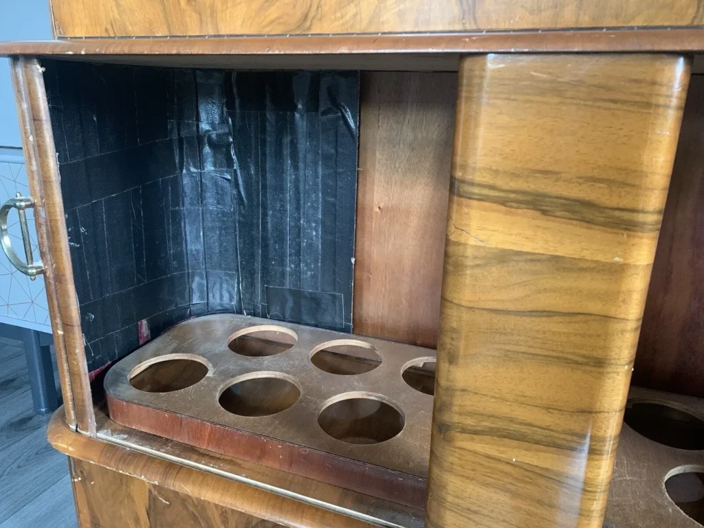 A wooden cabinet with an interior featuring multiple circular holes on a lower shelf, likely for holding bottles. The cabinet has a brass handle on the left side and a section lined with black tape on the inner back wall. The exterior wood has a glossy finish.