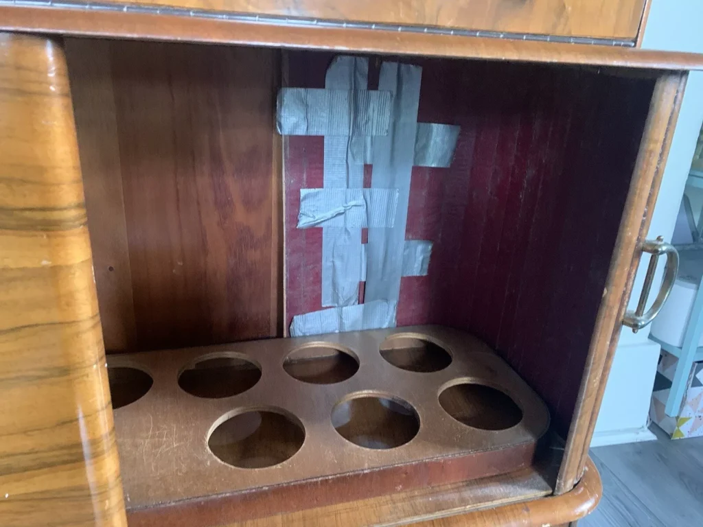 A wooden cabinet with a rectangular opening sits empty, save for several strips of duct tape covering the back panel. The shelf inside has circular cutouts, possibly for storing bottles or similar items. The wood has a polished finish, and there's a handle on the right side.