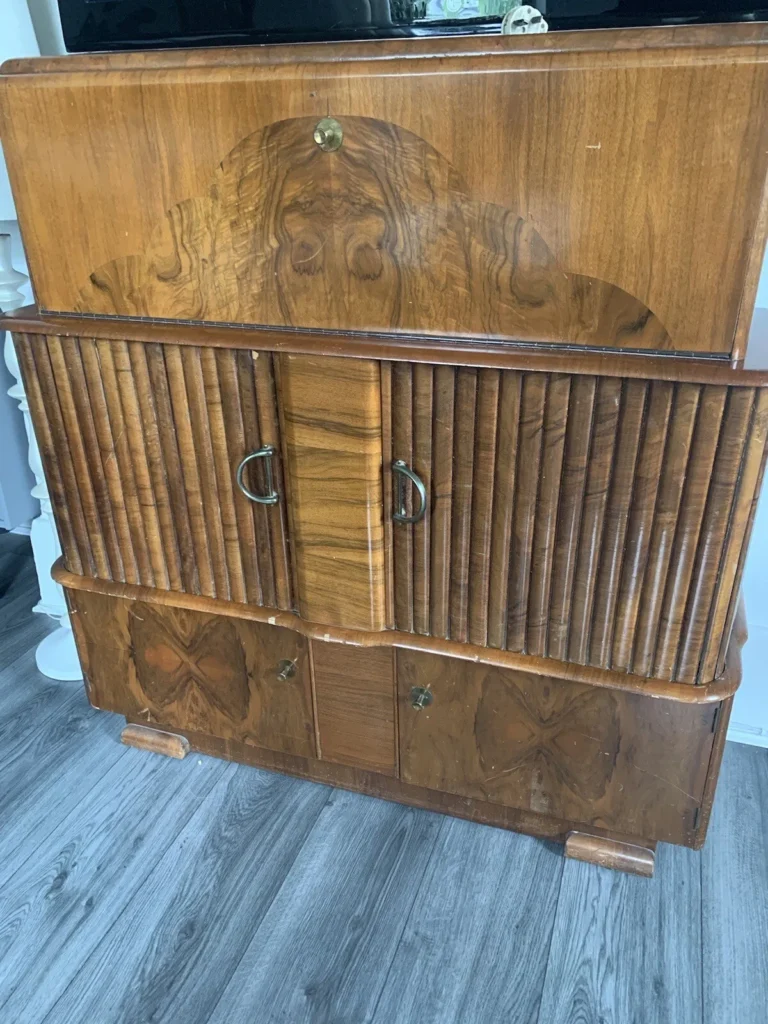 A vintage wooden cabinet with a decorative inlay on the top section, featuring two circular brass handles on the middle section with vertical grooves, and additional storage space on the bottom with two square brass handles. The cabinet stands on a gray wooden floor.