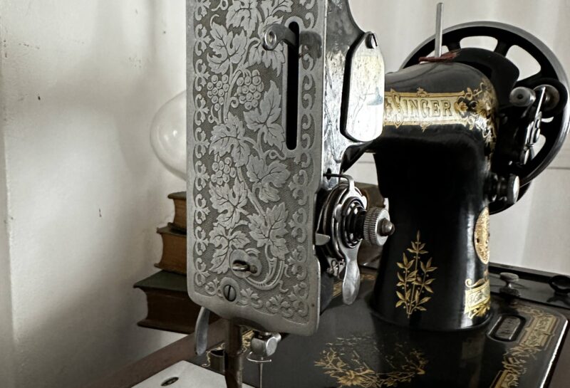 A close-up of an antique sewing machine with intricate decorative floral patterns on its metal parts. The machine includes a large handwheel and a needle mechanism. The sewing machine sits on a wooden table with additional floral designs. A white curtain is in the background.