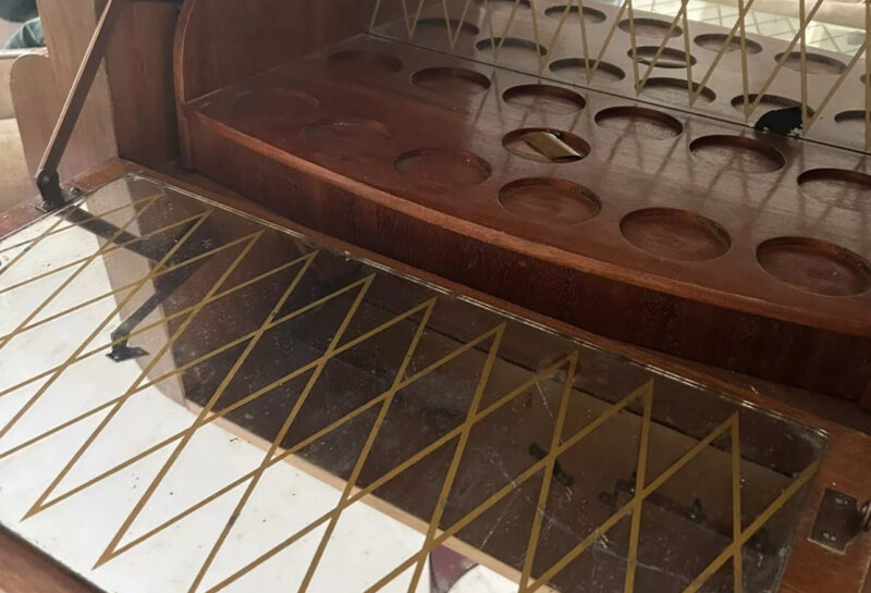 A wooden cabinet with a glass front showcasing an intricate design. Inside, there are multiple circular indentations and a mirrored surface with a geometric pattern. The cabinet appears to have metal hinges and supports.