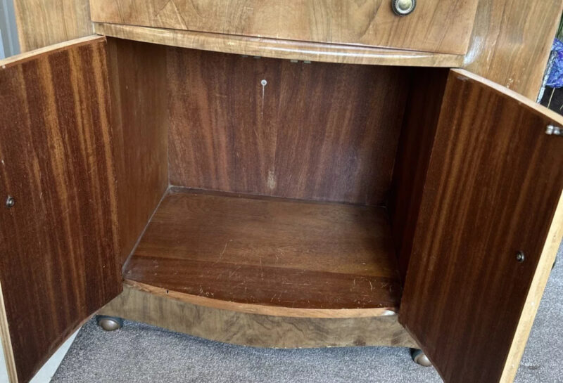 A wooden cabinet with two open doors on the lower half, revealing an empty storage space. Above the doors is a drawer with two round knobs. The cabinet stands on a carpeted floor, and some miscellaneous items, including a piece of paper and a white wire, are visible at the bottom right corner.