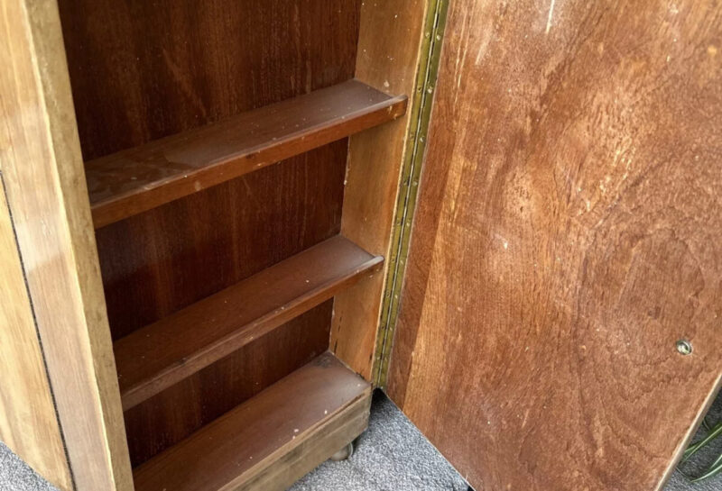 A wooden cabinet with an open door revealing two interior shelves. The cabinet has a natural wood finish, and it sits on a grey carpeted floor. A dark patterned blanket is draped over a chair in the background.