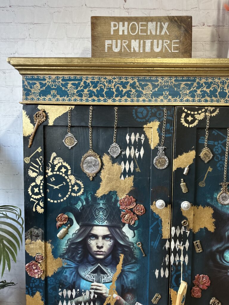 A painted cabinet with intricate designs including a woman holding cards, clocks, flowers, and keys. Gold leaf accents and various hanging ornaments embellish the piece. A sign on top reads "Phoenix Furniture.