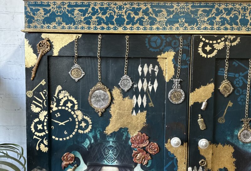 A painted cabinet with intricate designs including a woman holding cards, clocks, flowers, and keys. Gold leaf accents and various hanging ornaments embellish the piece. A sign on top reads "Phoenix Furniture.