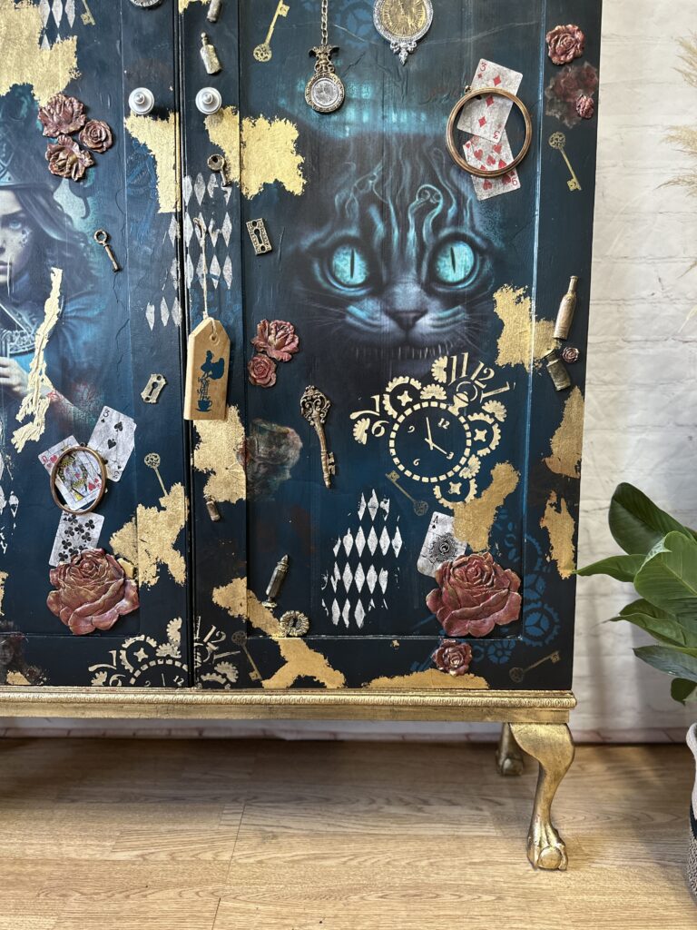 An ornate cabinet features an Alice in Wonderland theme with a large image of the Cheshire Cat with glowing blue eyes. Surrounding the cat are playing cards, clocks, keys, and roses, all set against a dark background with gold accents.