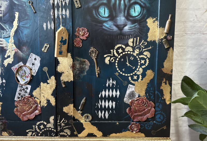 An ornate cabinet features an Alice in Wonderland theme with a large image of the Cheshire Cat with glowing blue eyes. Surrounding the cat are playing cards, clocks, keys, and roses, all set against a dark background with gold accents.