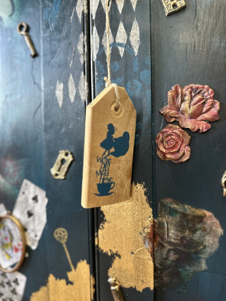 A wooden tag with the silhouette of a girl holding a teacup and the text "We’re All Mad Here" hangs on a string. Behind it is a dark-colored surface with artistic decor, including roses, playing cards, and keys. Some areas are painted gold.