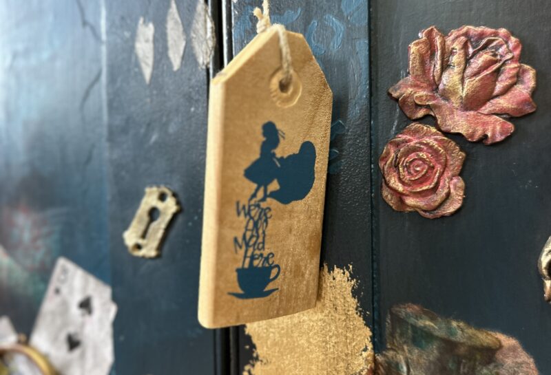 A wooden tag with the silhouette of a girl holding a teacup and the text "We’re All Mad Here" hangs on a string. Behind it is a dark-colored surface with artistic decor, including roses, playing cards, and keys. Some areas are painted gold.