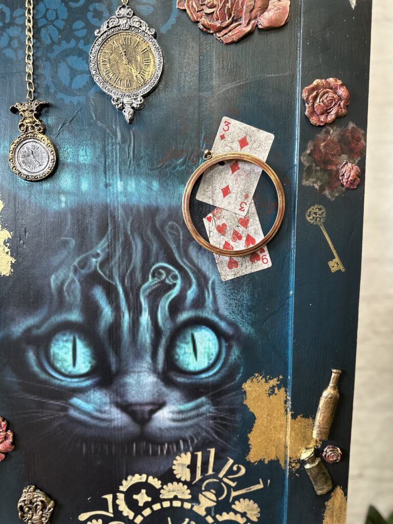 An intricately decorated door featuring a large, glowing blue-eyed cat's face, antique pocket watches, playing cards, keys, and roses. The golden Roman numerals and detailed artwork evoke a whimsical, fantasy theme.