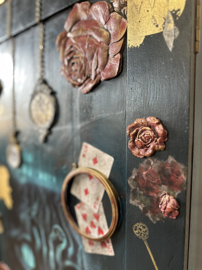 A close-up of a dark green box adorned with decorative elements: 3D floral designs, playing cards, embroidery hoop, vintage-style lockets, and gold painted accents. The overall aesthetic is eclectic and artistic, with a mix of textures and patterns.