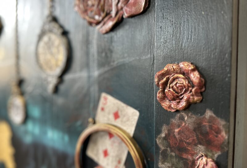 A close-up of a dark green box adorned with decorative elements: 3D floral designs, playing cards, embroidery hoop, vintage-style lockets, and gold painted accents. The overall aesthetic is eclectic and artistic, with a mix of textures and patterns.