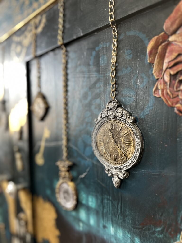 A close-up of an intricately designed antique pocket watch hanging by a chain against a dark, textured background with another pocket watch and a painted rose visible in the background. The watch features ornate details and Roman numerals on its face.