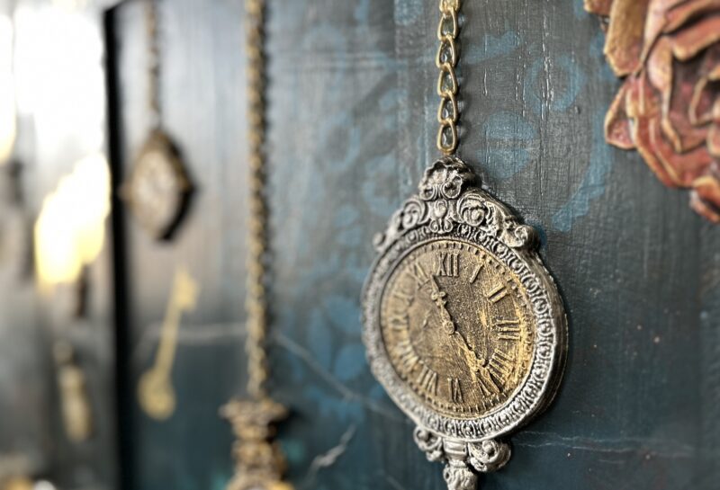 A close-up of an intricately designed antique pocket watch hanging by a chain against a dark, textured background with another pocket watch and a painted rose visible in the background. The watch features ornate details and Roman numerals on its face.