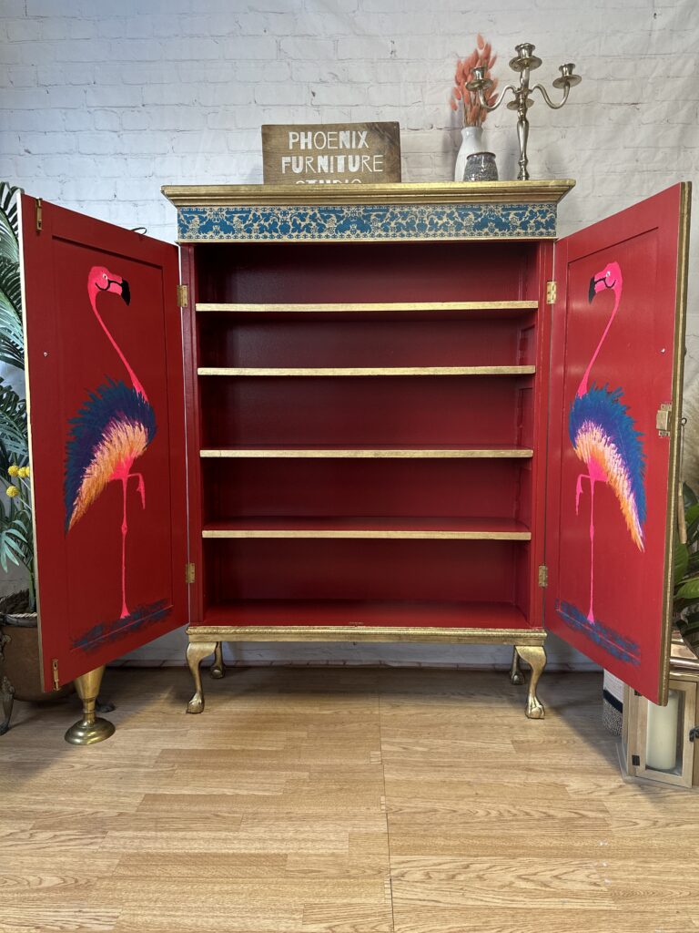 An open wooden cabinet with four shelves, painted red inside. The cabinet doors feature colorful flamingo artwork. The exterior is accented with gold and blue details. The sign "Phoenix Furniture" is placed on top. Plants and a candle stand surround the cabinet.