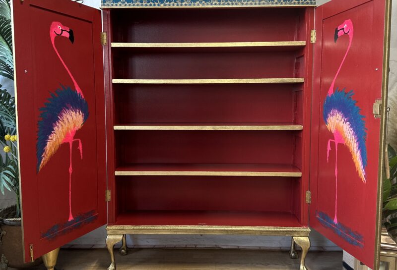 An open wooden cabinet with four shelves, painted red inside. The cabinet doors feature colorful flamingo artwork. The exterior is accented with gold and blue details. The sign "Phoenix Furniture" is placed on top. Plants and a candle stand surround the cabinet.