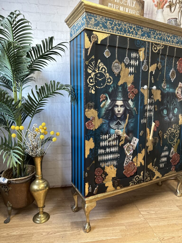 A decorative cabinet with intricate designs, featuring an illustrated woman in a top hat, playing cards, and roses. The cabinet has blue, gold, and black details. To the left is a potted palm plant and a brass vase with dried flowers. The setup is on a wooden floor.