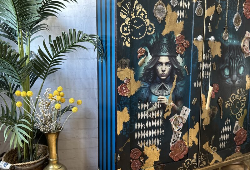 A decorative cabinet with intricate designs, featuring an illustrated woman in a top hat, playing cards, and roses. The cabinet has blue, gold, and black details. To the left is a potted palm plant and a brass vase with dried flowers. The setup is on a wooden floor.