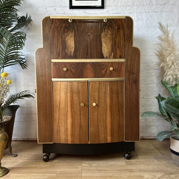 A wooden cabinet with an art deco design featuring gold handles and hinges stands on a light wood floor. Above it hangs a framed black-and-white line drawing. The scene is adorned with potted plants, including palms and ornamental grasses, against a white brick wall.