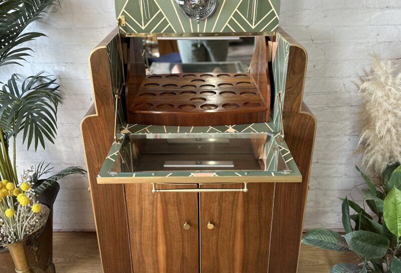 A vintage wooden cabinet with an upper section featuring geometric-patterned green and beige accents and an open shelf with circular cutouts. Below, double doors and brass handles provide storage. The cabinet is decorated with plants and set in a room with a white brick wall.