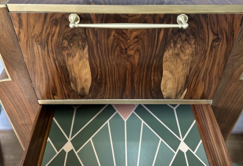 A wooden drawer unit with brass handles and trim is shown. The lower drawer is open, revealing a geometric design on the interior in green and pink tones. The unit is positioned against a white brick wall, and the floor is wooden.