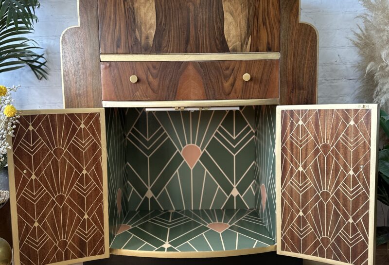 A vintage-style wooden bar cabinet with art deco design elements. The cabinet has an upper compartment, a drawer, and two lower doors with geometric patterns, filled with green and gold. A framed abstract line drawing hangs on a white brick wall in the background.