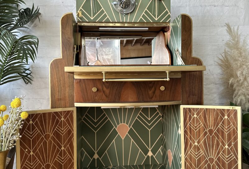 A vintage wooden bar cabinet with an open top showcasing glassware and a bottom section with open doors. The interior features a green and gold geometric design. The cabinet stands on castor wheels and has decorative plants and a framed artwork nearby.