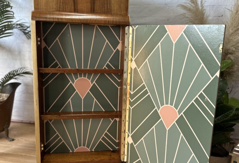 A vintage wooden cabinet with an open door reveals three interior shelves and a decorative green and pink geometric pattern on the back panel. The cabinet is set against a white brick wall, with some plants and a framed picture in the background.