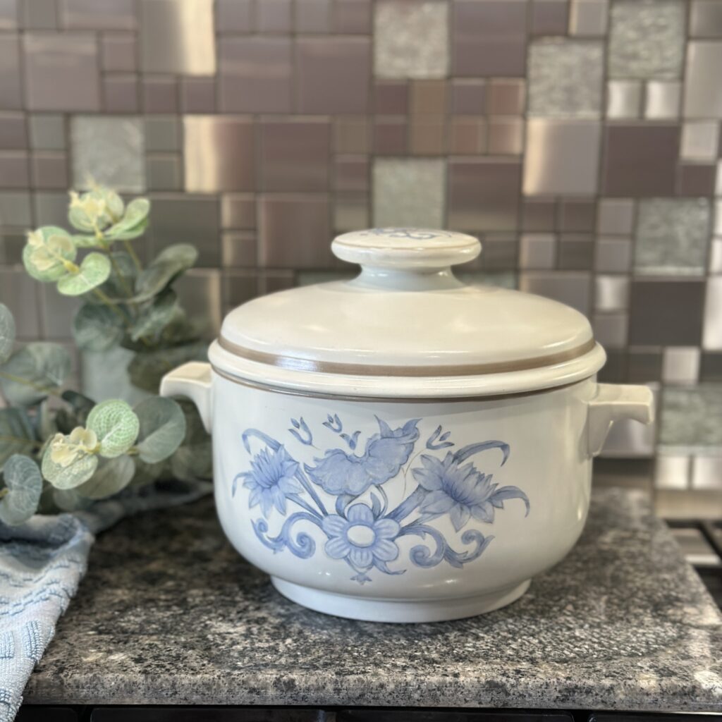 A white ceramic pot with a lid, featuring blue floral designs, sits on a granite countertop. A green leafy plant is placed beside it, and a tiled backsplash with varied shades of brown and gray is in the background.