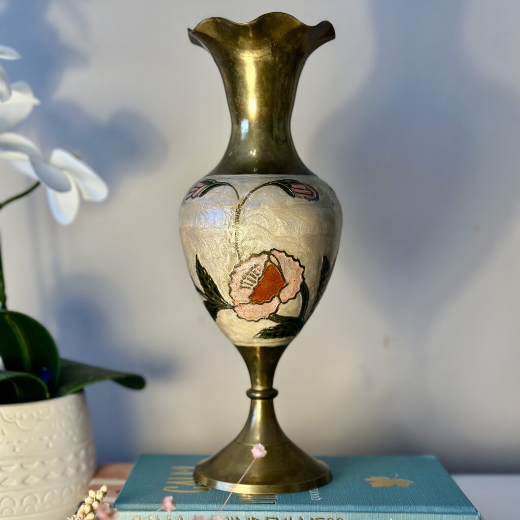 A decorative brass vase with a floral design stands atop a blue book. Its base and rim are polished brass, while the middle features painted flowers. An orchid plant and a beaded necklace are partially visible to the left.