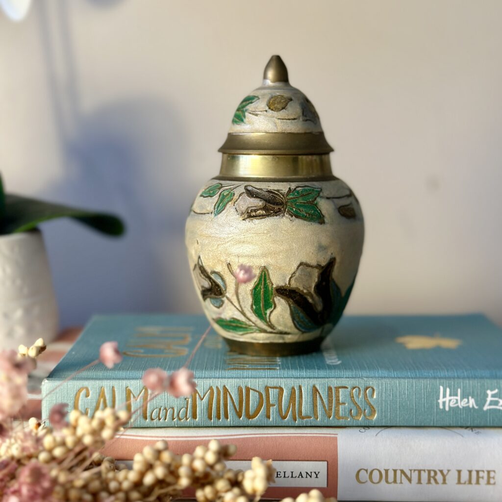 A decorative urn with leaf and flower motifs sits on two stacked books titled "Calm and Mindfulness" and "Country Life." In the foreground, there are dried flowers. A blurred plant is in the background on the left.