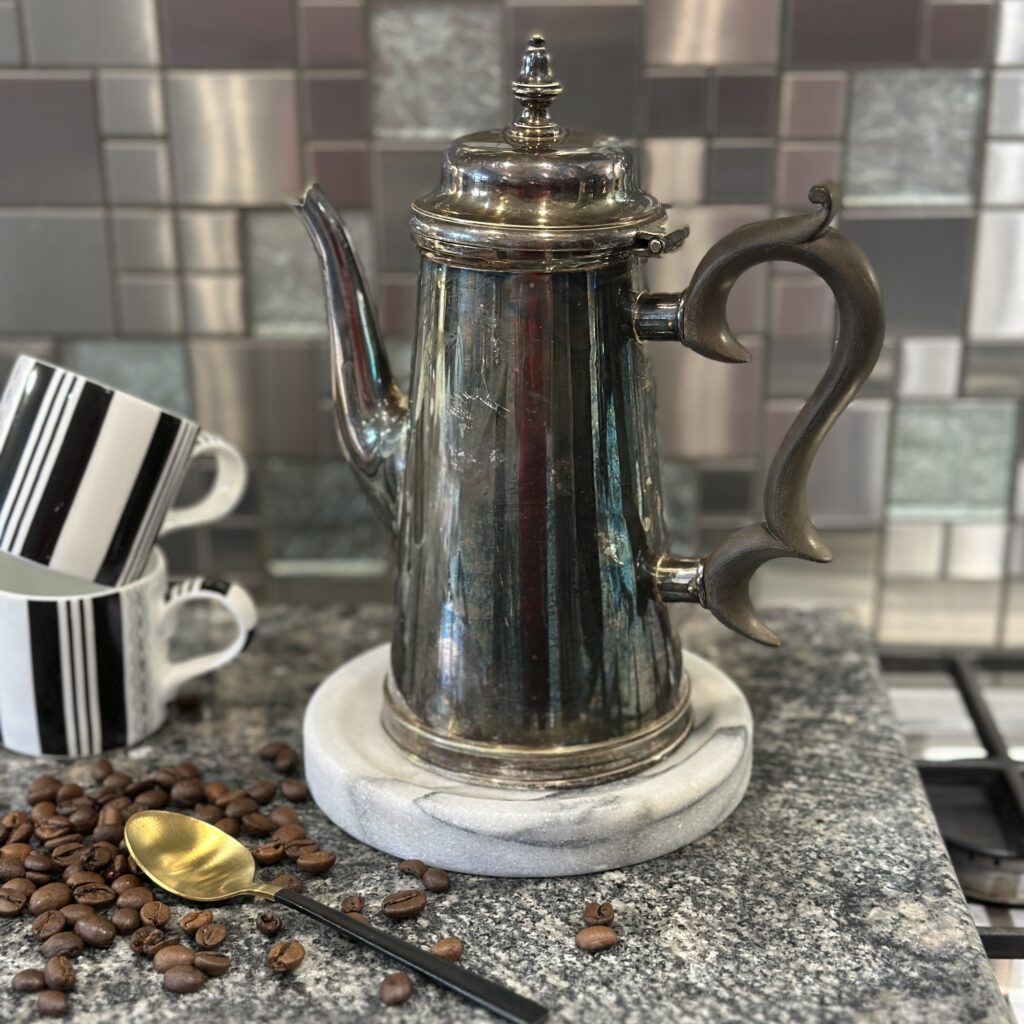 A vintage silver coffee pot with a curved spout and handle sits on a circular marble coaster. Two black-and-white striped mugs and scattered coffee beans with a small golden spoon accompany the pot on a granite countertop. A mosaic tiled backsplash is visible.