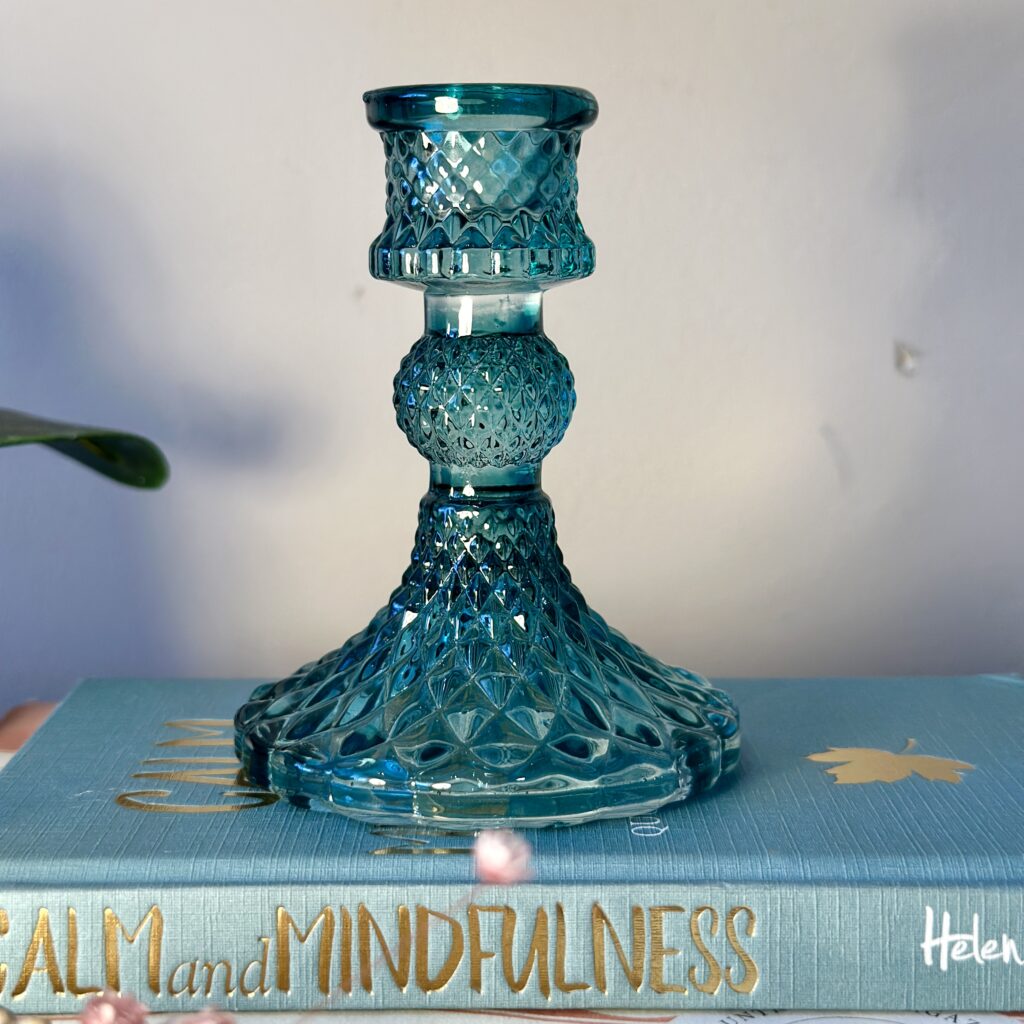 A blue glass candlestick holder with intricate patterns sits on a hardcover book titled "Calm and Mindfulness." A small flower is in the foreground, and a plant leaf peeks in from the left.