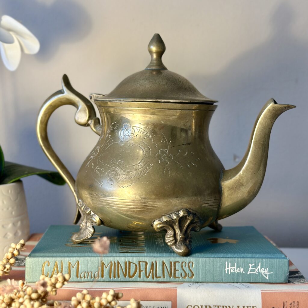 A brass teapot with floral engravings sits atop three stacked books, titled "CALM and MINDFULNESS" and "COUNTRY LIVING." A white orchid and a sprig of small white flowers accompany them, creating a serene still life.