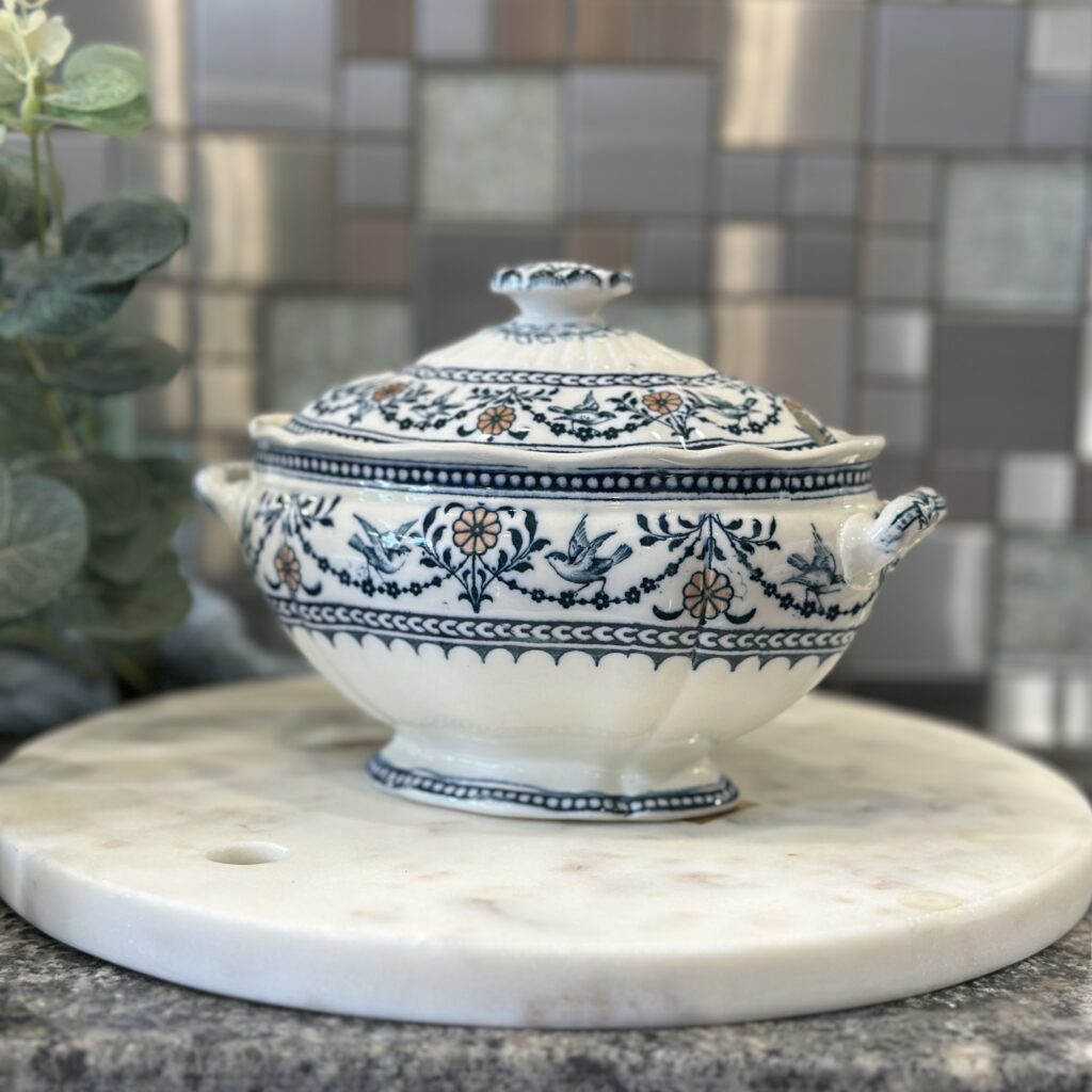 A decorative ceramic soup tureen with floral and leaf patterns sits on a round marble board. The background features a tiled wall in various shades of grey.