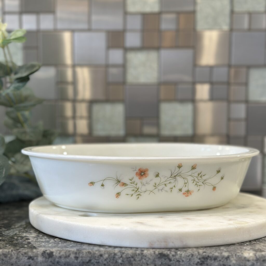 A white dish with floral details rests on a round marble tray. The background features a mosaic tile wall in shades of gray and beige, with green foliage to the side.