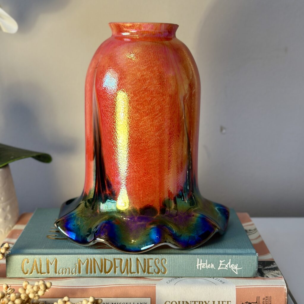 A vibrant, glossy orange and multicolored glass vase sits on a stack of books titled "Calm and Mindfulness" and "Country Life." The vase has a flowing design, creating a striking and artistic display on the bookshelf.