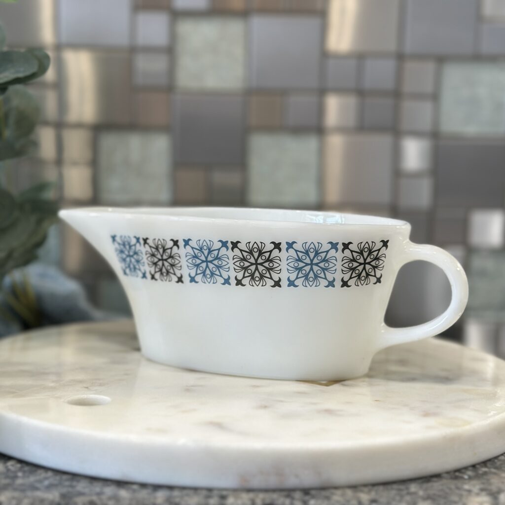 A white ceramic gravy boat with a handle and a decorative blue and black floral pattern sits on a round marble platform. The background features a mosaic tile wall in various shades of grey and beige.