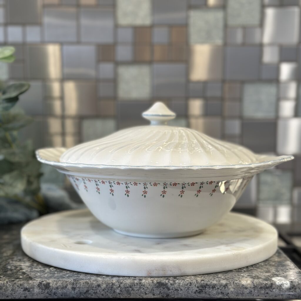 A decorative ceramic soup tureen with a lid sits on a round marble base. The tureen features delicate floral patterns and is placed against a mosaic tile backsplash. Subtle greenery is visible on the left side.