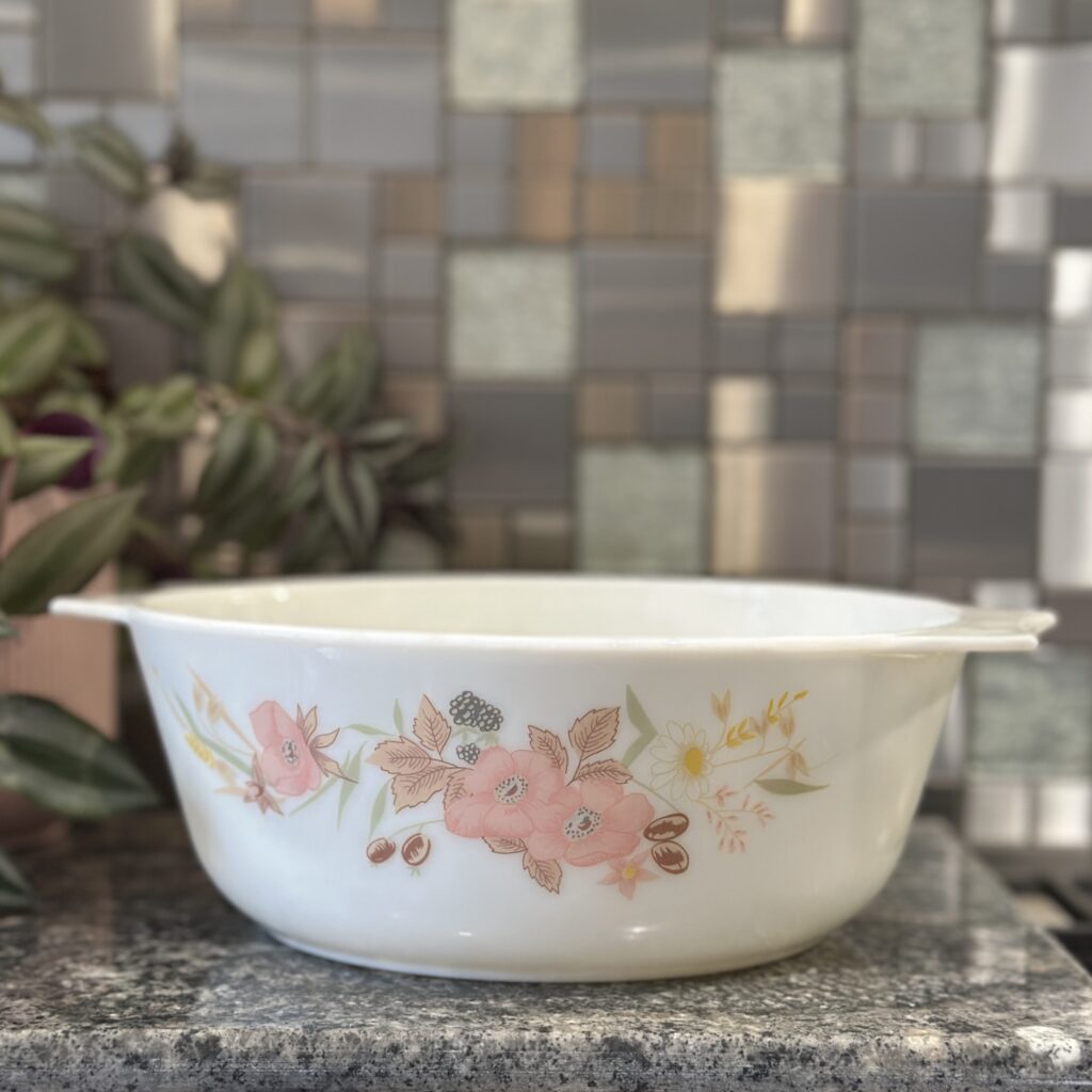 A vintage white mixing bowl with pink floral patterns sits on a countertop. Behind it, there's a plant with green and purple leaves on the left, and a multicolored tiled backsplash in shades of gray, blue, and beige.