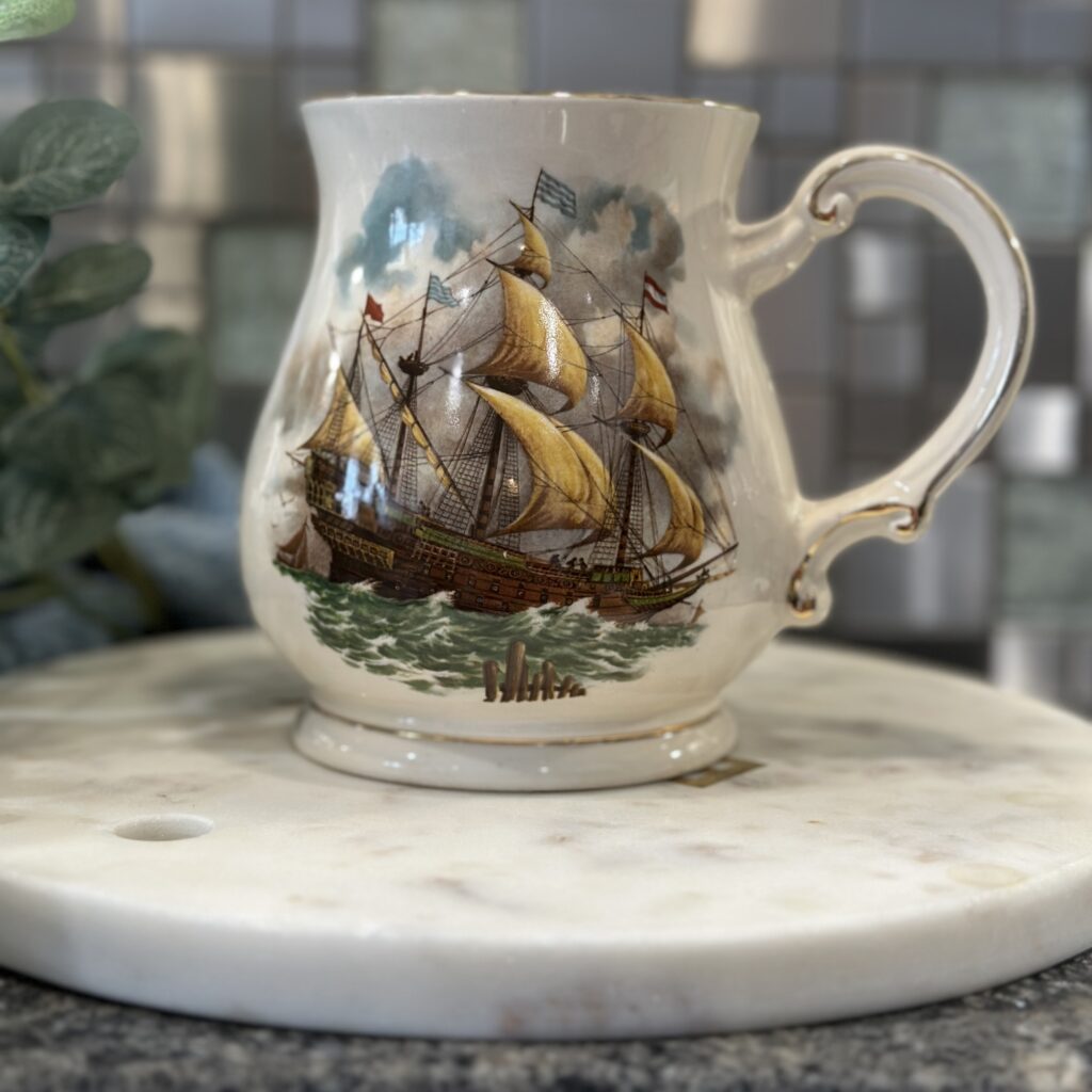 A decorative ceramic mug featuring a vintage sailing ship on the ocean, placed on a round marble coaster. Green foliage is visible in the background.