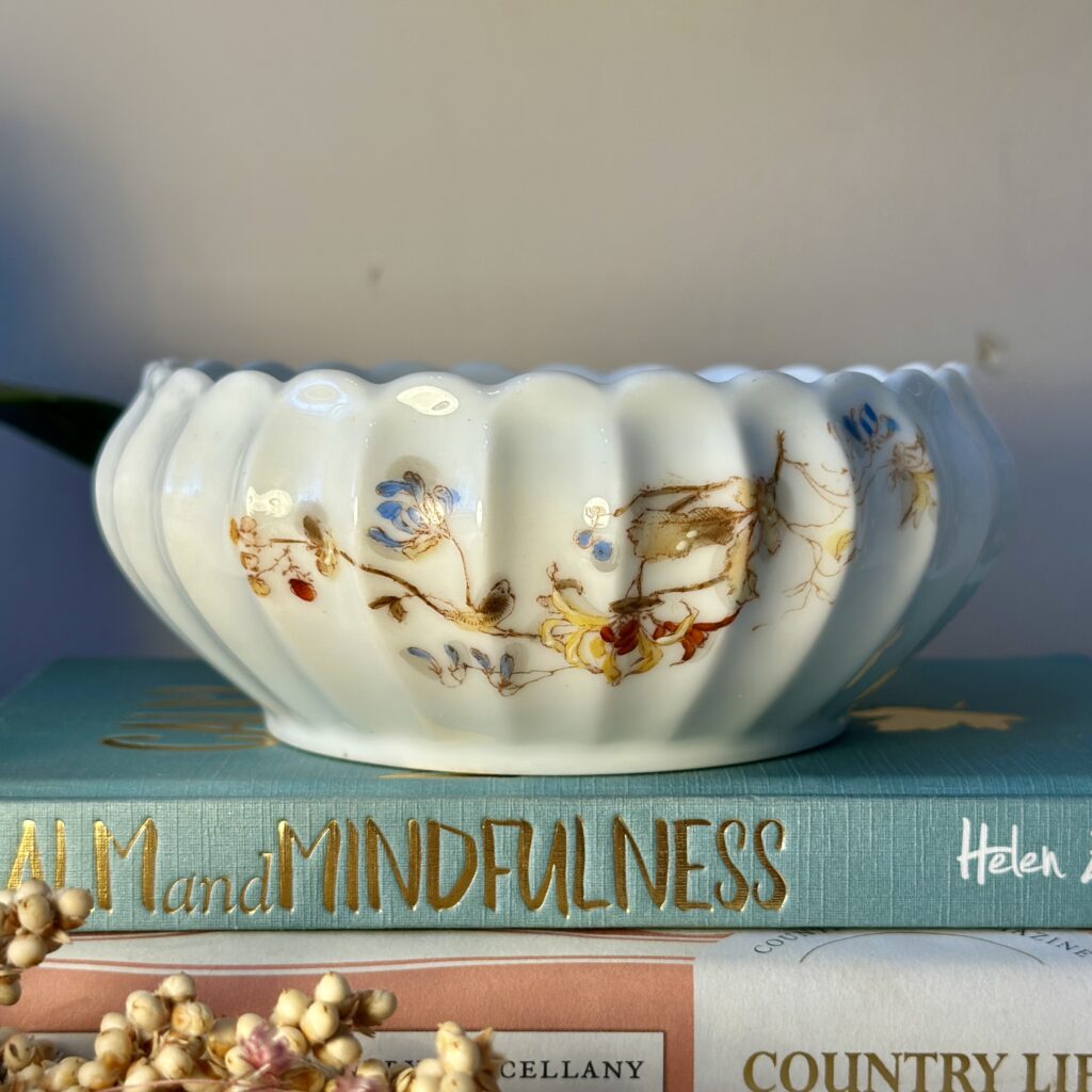 A white, scalloped ceramic bowl with delicate floral designs in blue, yellow, and red sits on a stack of hardcover books. The titles on the books are partially visible, and a small sprig of dried flowers rests in the foreground.