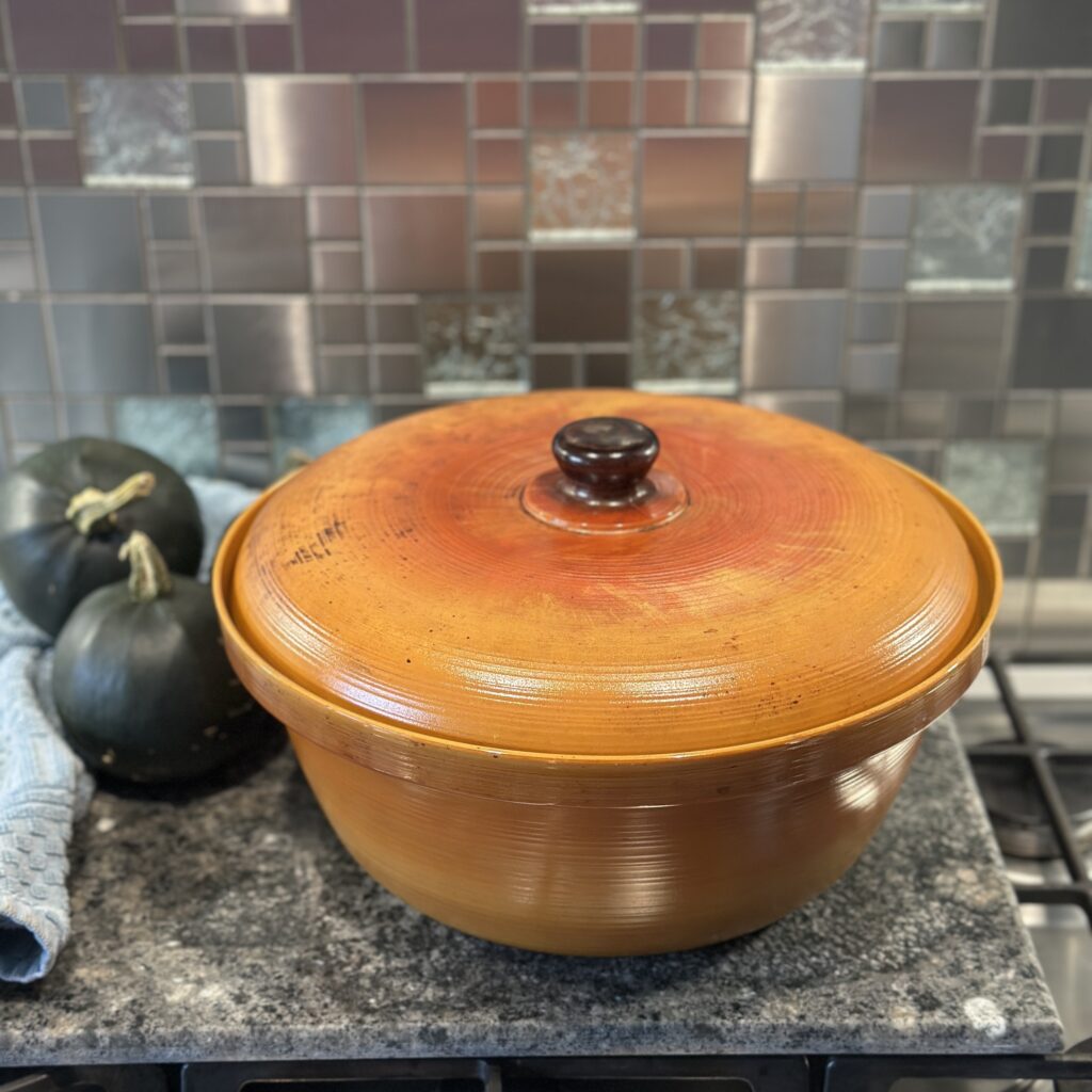 A large, round clay pot with an orange lid sits on a stove. Two dark green squash are placed beside the pot. The background features a metallic mosaic tile backsplash, and a light blue dish towel is partially visible.