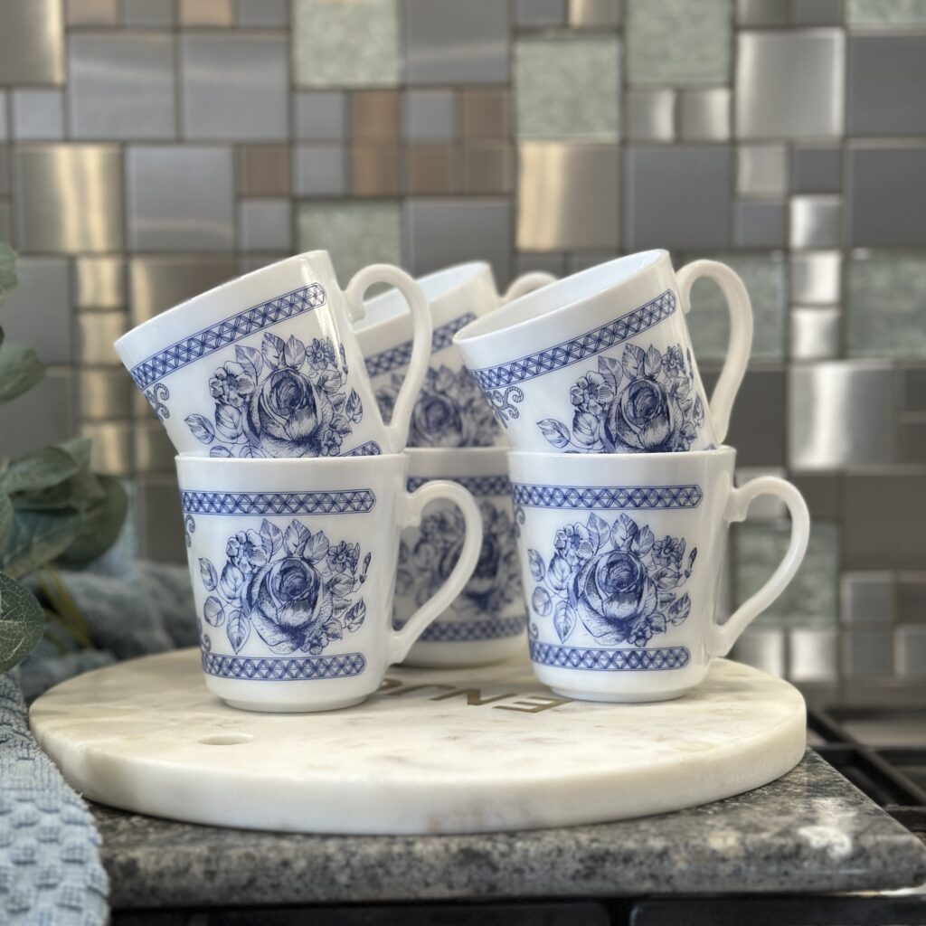 Six white mugs with blue floral patterns are stacked on a white round marble slab. The background features a metallic and tiled backsplash.