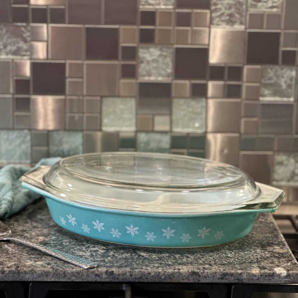 A vintage teal casserole dish with a clear glass lid, featuring a white snowflake pattern, sits on a granite countertop. The background features a mosaic tile backsplash in shades of grey and beige. A silver spoon lies nearby.