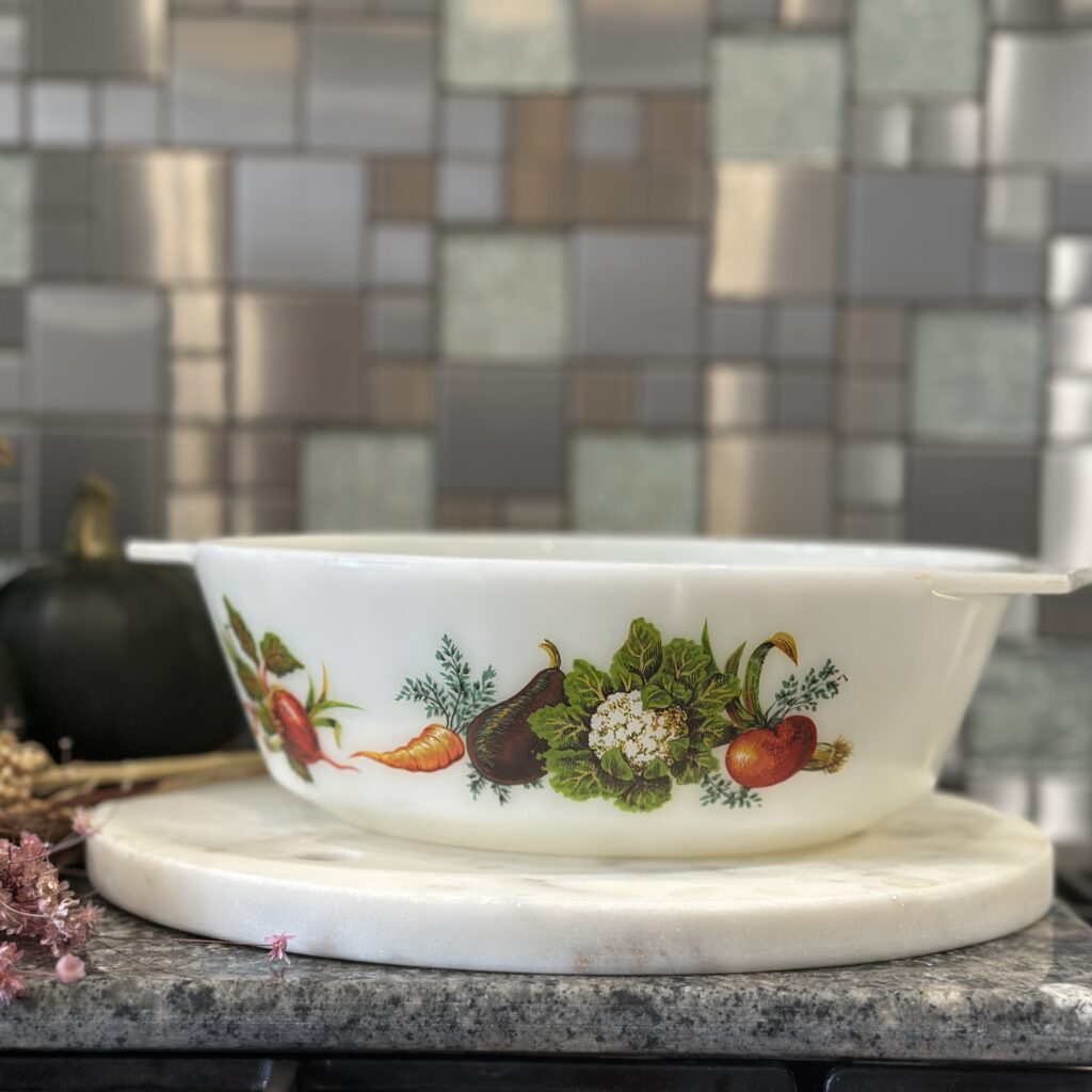 A white vintage dish with colorful vegetable illustrations, including a carrot and beet, is placed on a round marble tray. The background features a mosaic tile wall in shades of gray and beige.