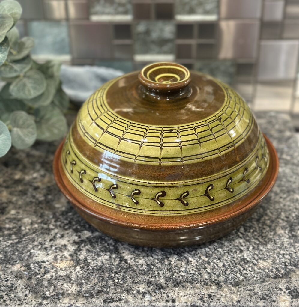 A decorative ceramic bowl with a matching lid sits on a granite countertop. The bowl features intricate green and brown patterns, with plant leaves visible in the background.