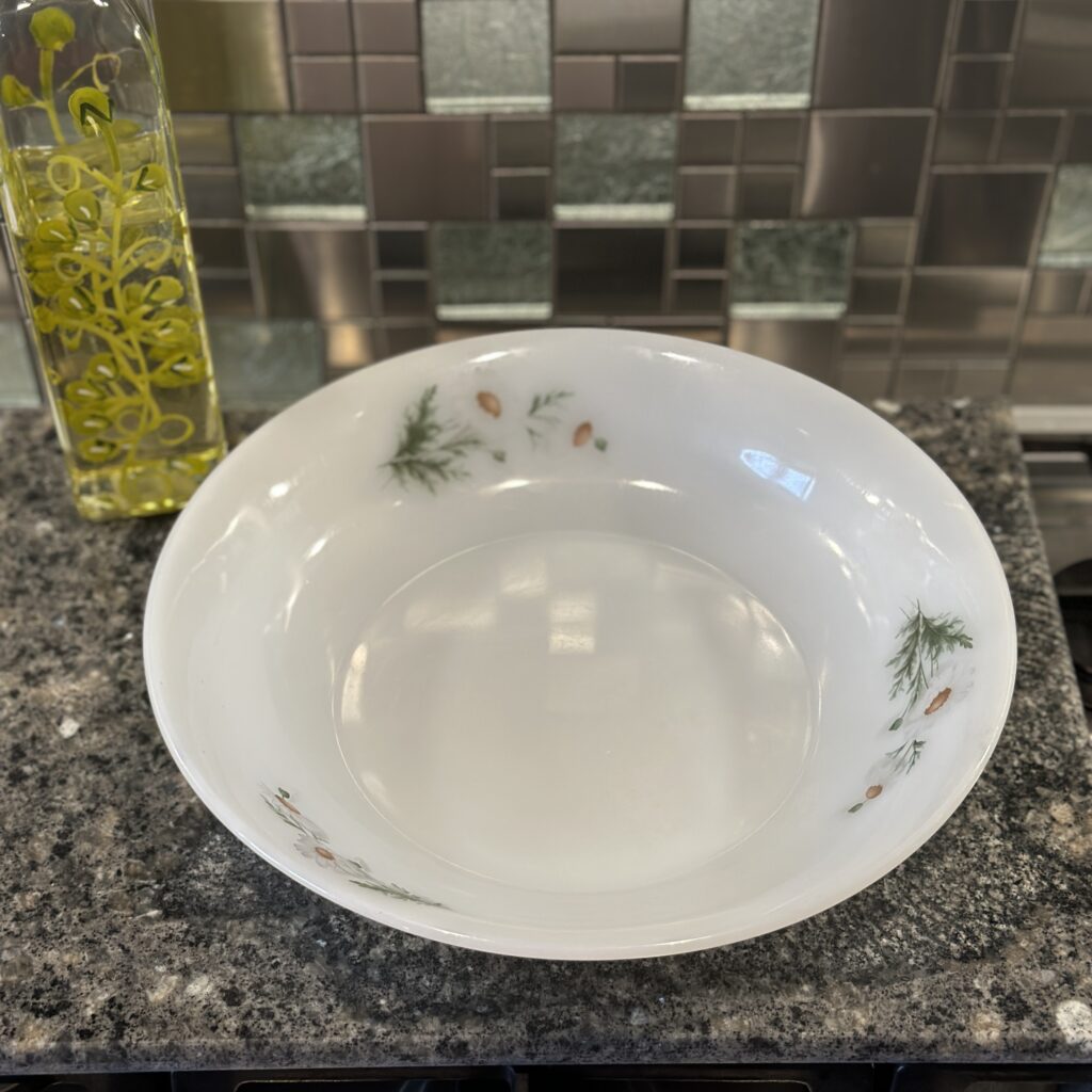 A large white glass salad bowl with a floral design sits on a granite countertop. Next to it is a tall bottle of olive oil. The background features a modern tiled kitchen backsplash.